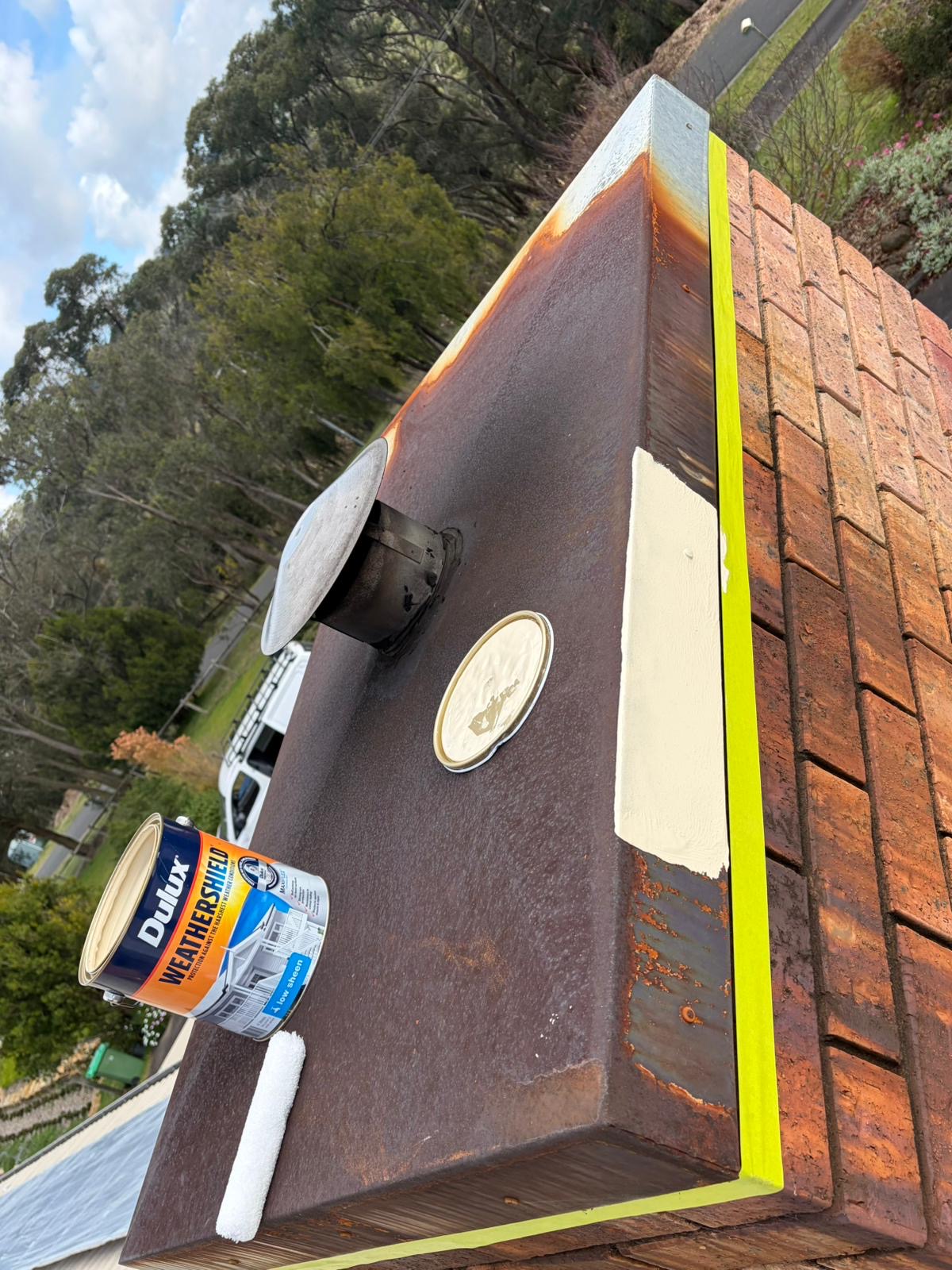 Rusted metal chimney chase cover being painted cream, with yellow tape along the brick and a Dulux Weathershield tin on the roof