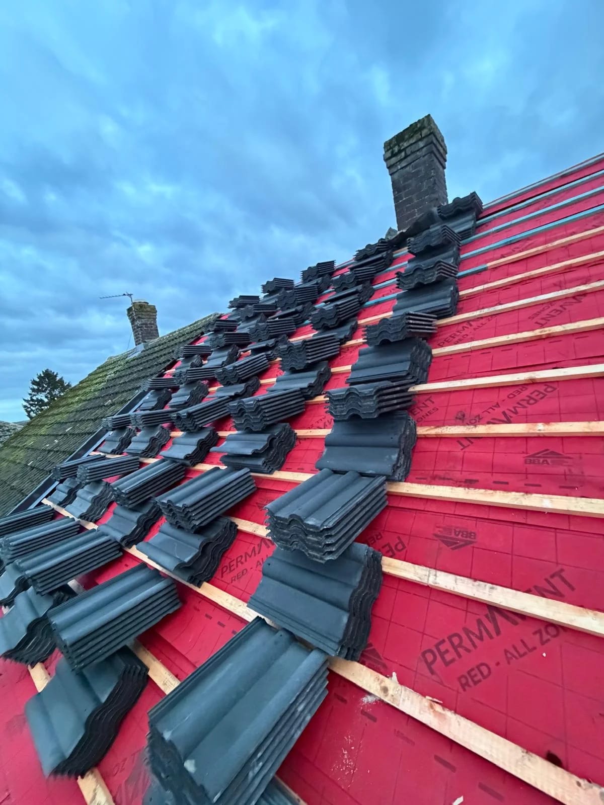 Red underlay and timber battens on a stripped roof, with stacks of dark tiles ready to lay